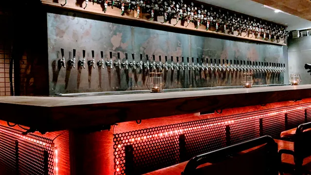 A metal wall with dozens of beer taps above a wooden counter and red lighting