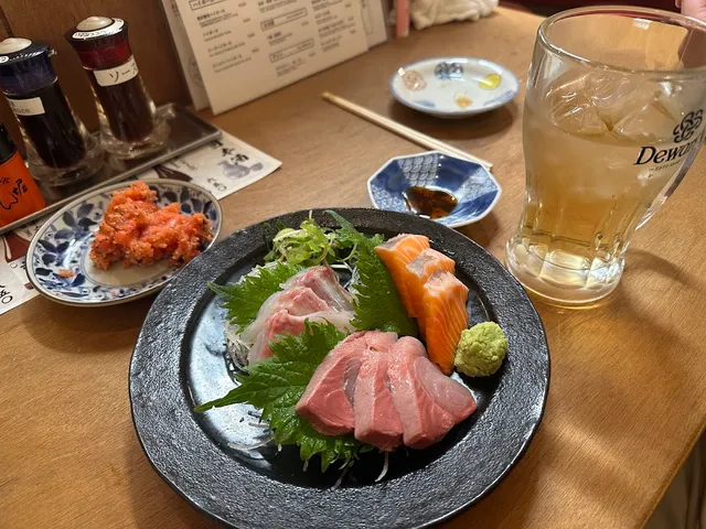 A plate of sashimi with shiso leaves and wasabi on a wooden counter, beside a glass of drink with ice