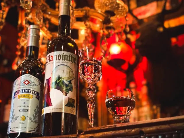 Two absinthe bottles and goblets on a counter, with a chandelier in red light behind