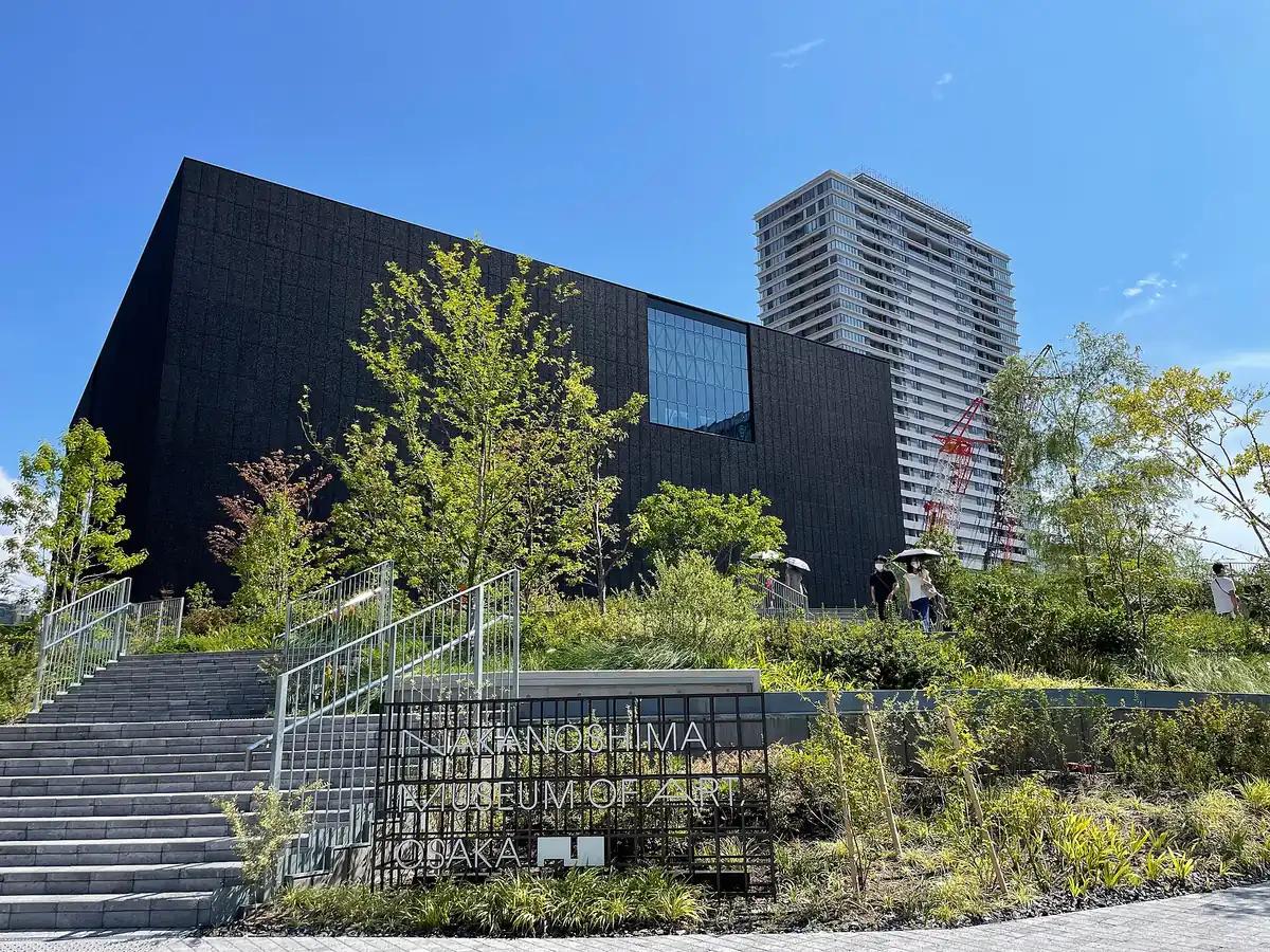 A black cube building with a large glass window, wide steps and planting in front under a blue sky