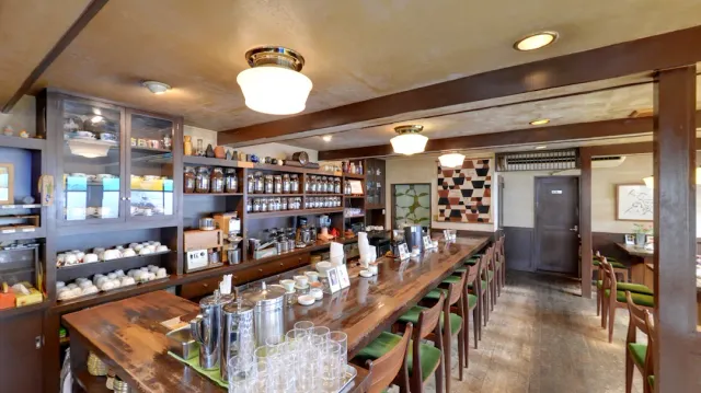 A long wooden counter with chairs, shelves of jars and wares and round ceiling lamps