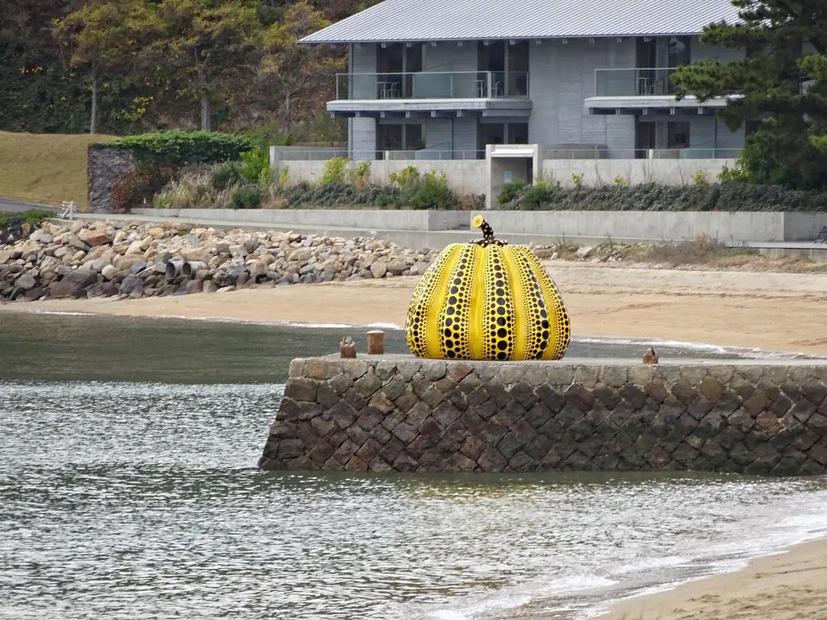 A yellow pumpkin sculpture with black dots on a stone pier at the edge of a quiet beach