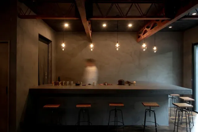 A long concrete counter with stools, hanging bulbs and a blurred figure behind it