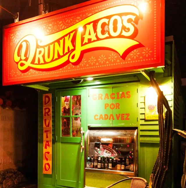 A green frontage at night with a large lit red sign, a service window with bottles and a cactus