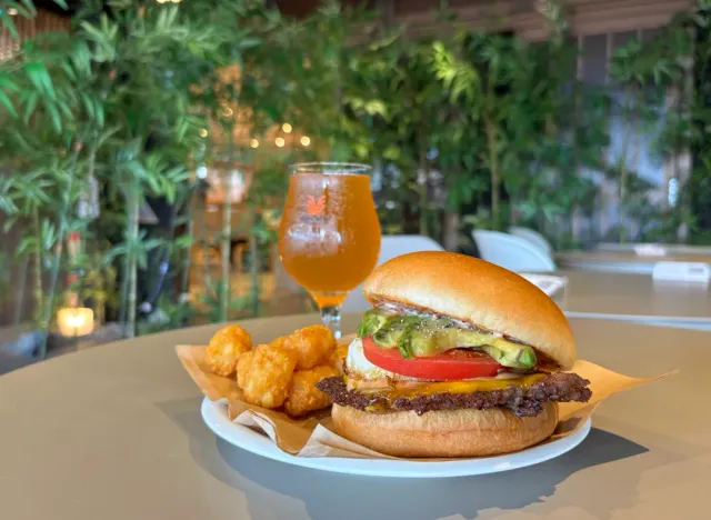 A burger in a bun with tomato and avocado beside potato bites and a glass of beer