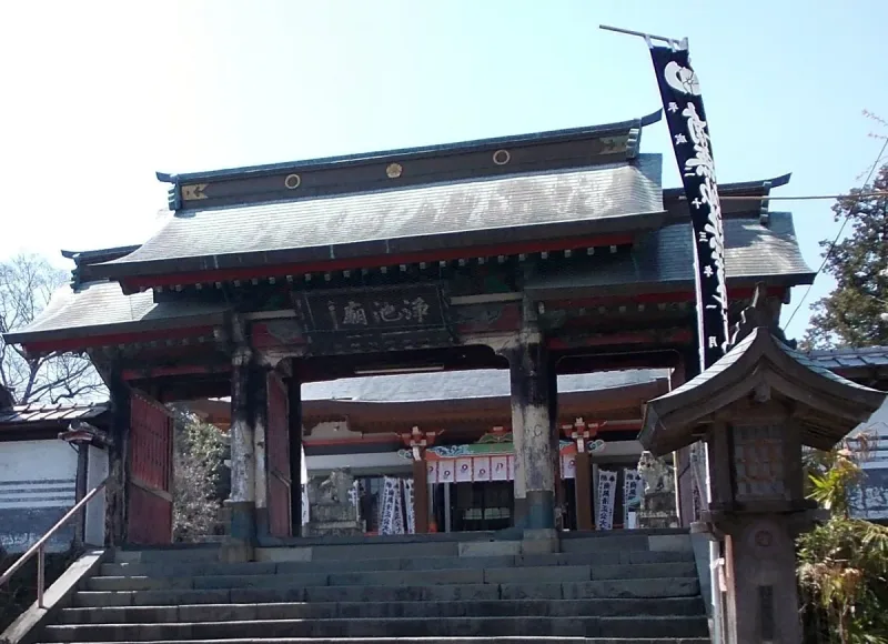 A wooden temple gate with a dark tiled roof and black banners, against a green hill
