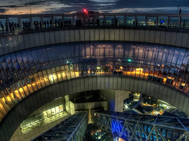An open ring shaped rooftop atop a tall building, people at the railing and city lights at night