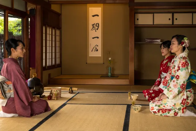 A tatami room with a calligraphy scroll, a woman beside a kettle facing two guests in kimono