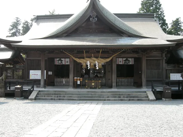 A wooden hall with a curved roof and a braided straw rope above the entrance, facing a gravel courtyard