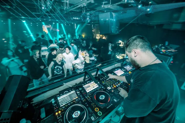 A DJ behind a mixing booth facing a dancing crowd in turquoise lights