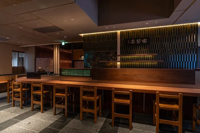 A long wooden counter with chairs, a lit slatted wall and a Japanese lettered sign on the wall