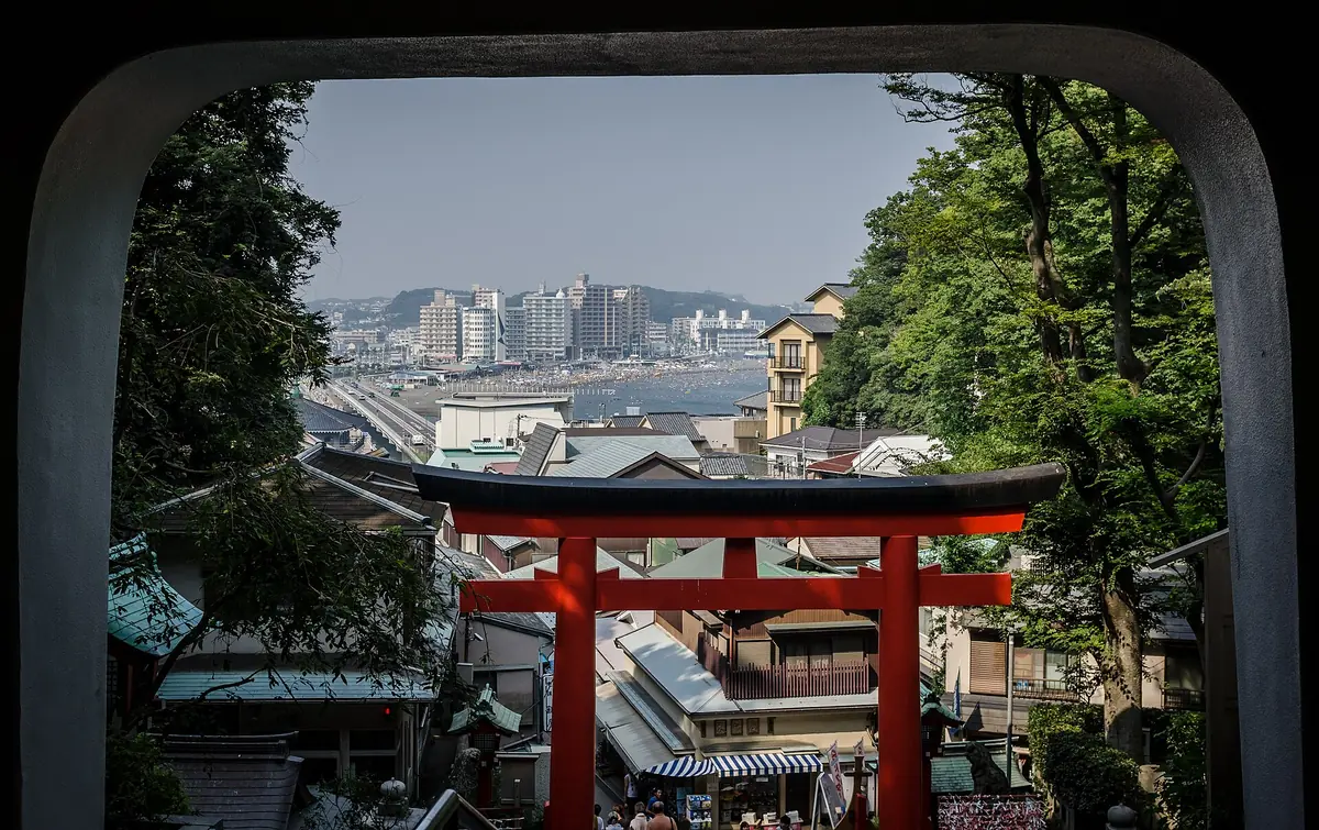 A red torii gate framed by a stone opening, with the town roofs and a coastal bay beyond