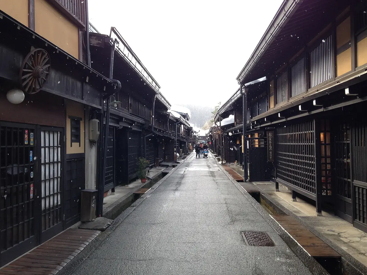 A street lined with traditional black wooden Edo-era merchant houses with lattice shutters in Takayama, light snow falling and people walking