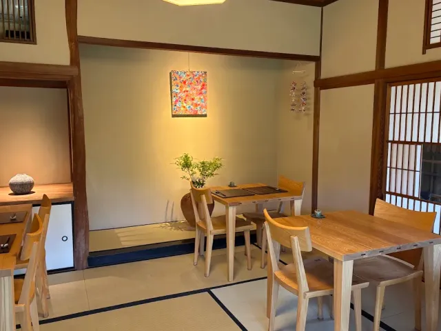 A tatami room with pale wooden tables and chairs, an alcove with a vase and a colourful picture