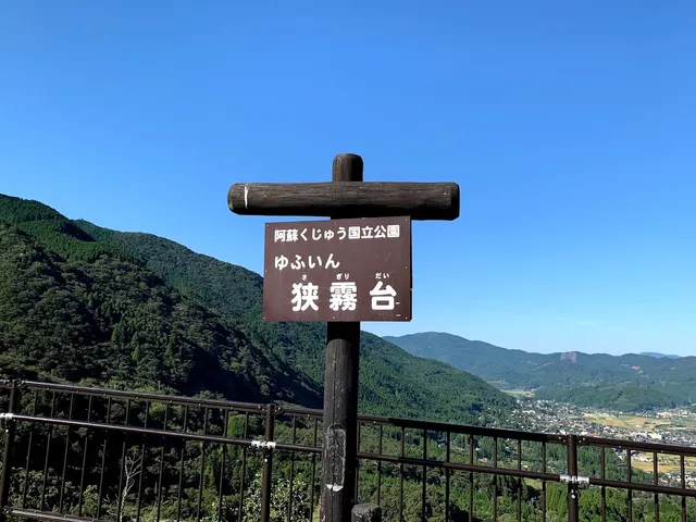 A wooden sign above a lookout railing, facing forested mountains and a valley under a blue sky