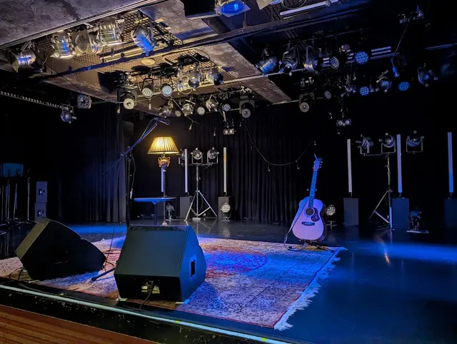 An empty stage with a rug, an acoustic guitar on a stand and blue stage lighting