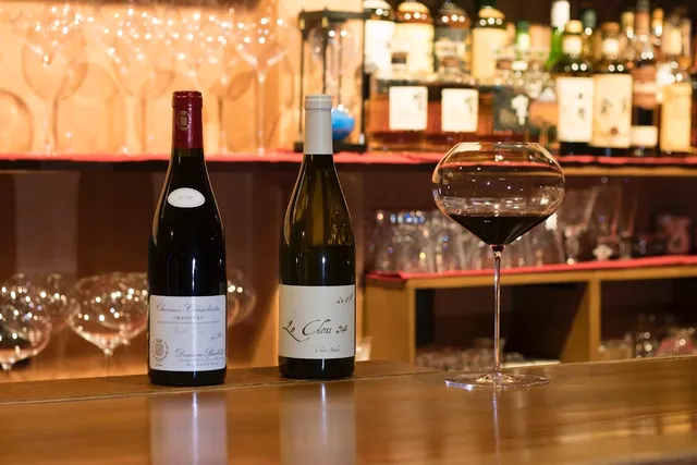 Two bottles of wine and a glass of red wine on a counter, in front of a shelf of bottles