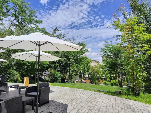 A paved terrace with white parasols and wicker armchairs facing a lawn and trees