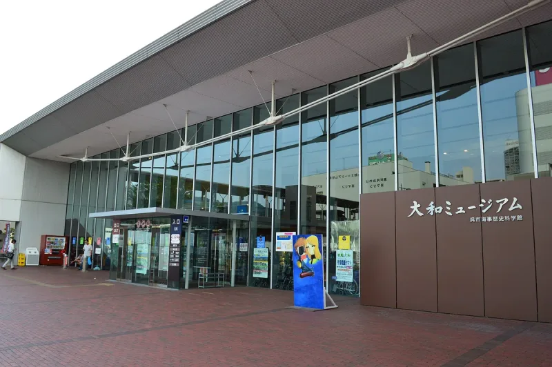 A glass clad museum entrance with a sign and posters, and a paved forecourt in front