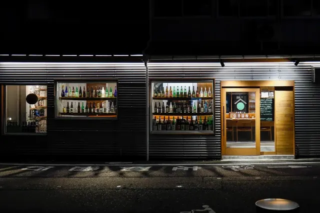 A corrugated metal frontage at night with lit windows holding rows of bottles, and a pale wooden door