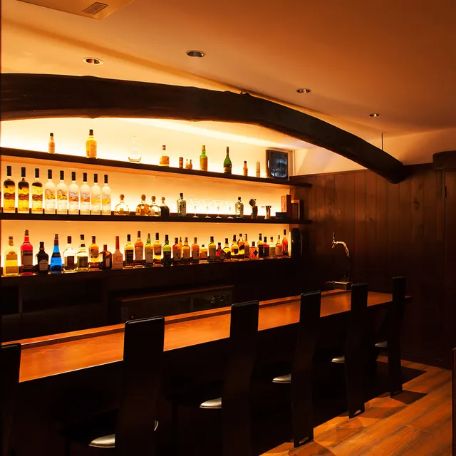 A wooden counter with high chairs facing lit bottle shelves and an arched beam