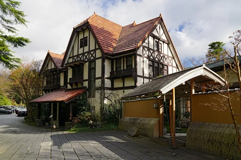 A European style timber framed house with a steep tiled roof, beside a Japanese entrance gate with an ochre earthen wall and bamboo fencing