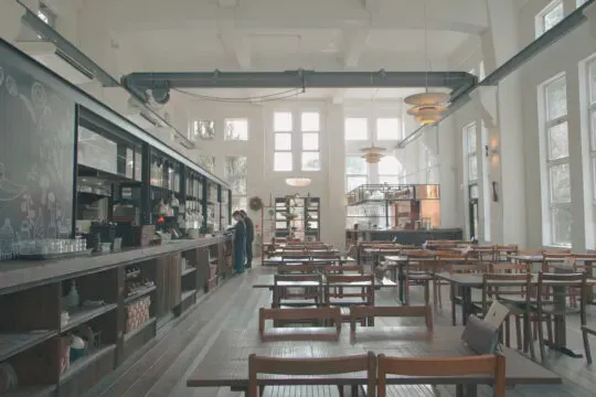 A tall bright hall with a long counter, wooden tables and chairs and tall windows