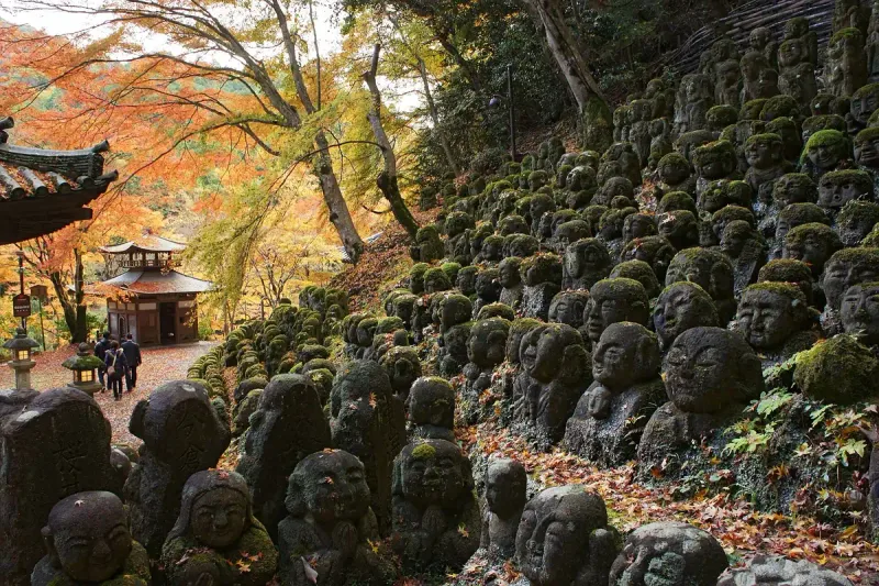 Hundreds of small moss covered stone statues on a slope in a temple courtyard, among autumn trees
