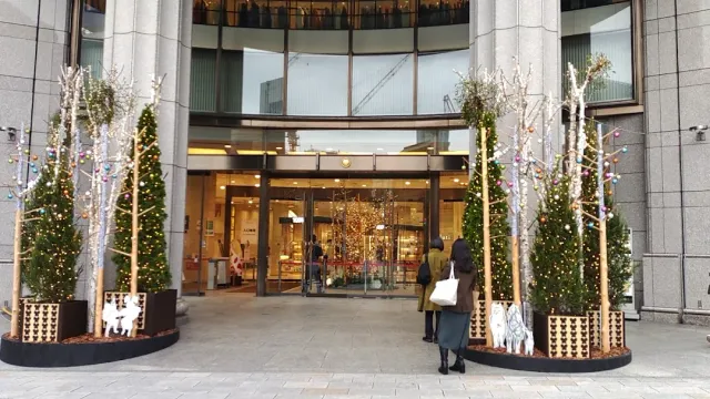 A lit glass entrance with fir trees decorated with baubles on both sides and customers walking in