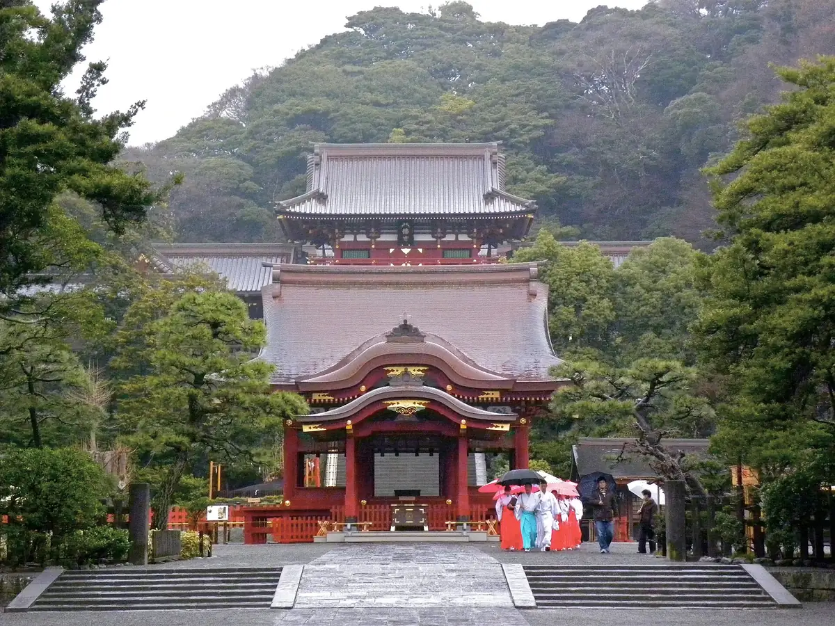 A red shrine hall at the top of wide stone steps, against a wooded hillside