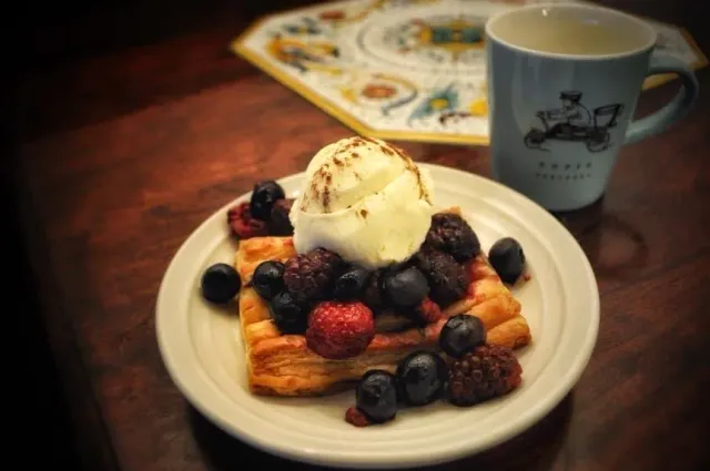 A puff pastry with berries and a scoop of white cream on a plate, beside a coffee mug
