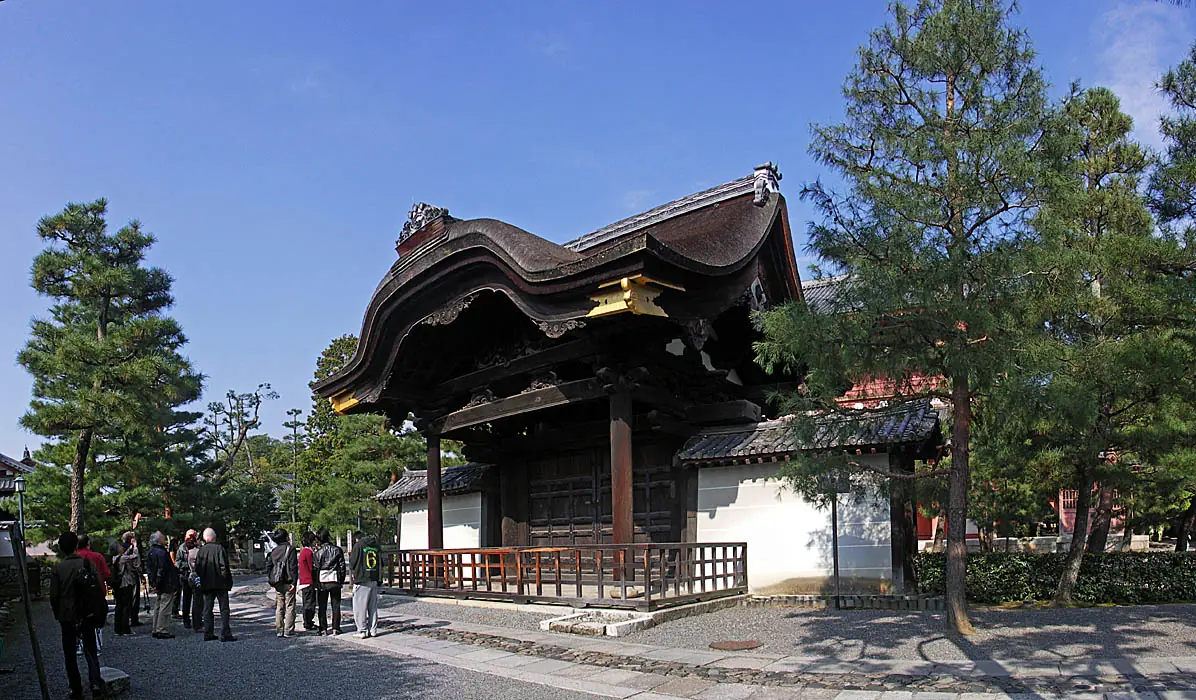 An old wooden gate with a dark undulating roof between pine trees under a blue sky