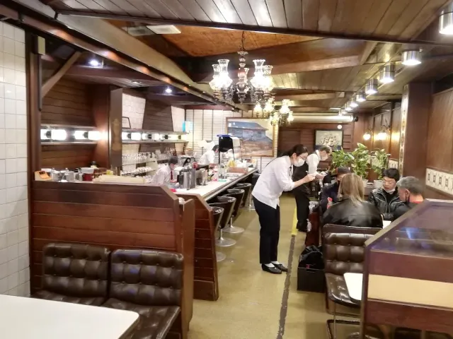 An old room with chandeliers, a long counter, brown leather benches and many customers