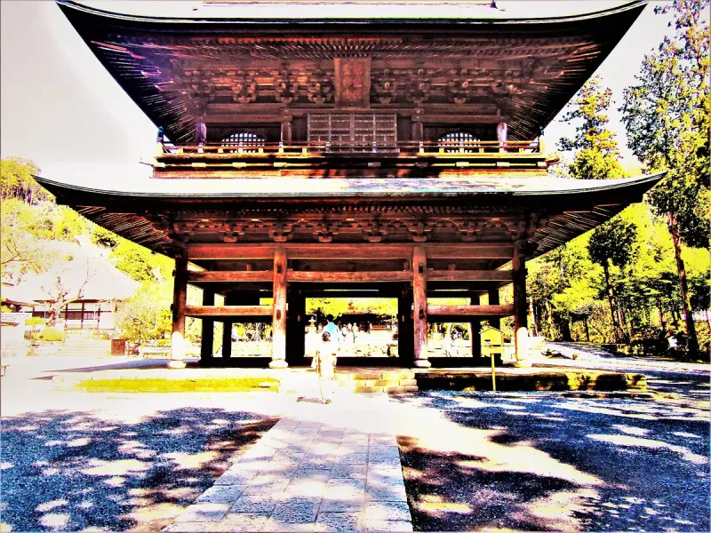 A two storey wooden temple gate with red beams and a tiled roof, among green trees