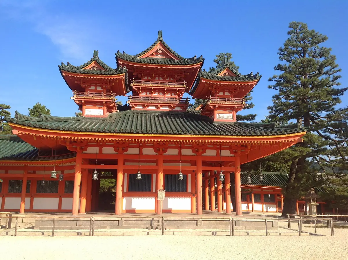 The vermilion entrance gate of Heian Jingu Shrine in Kyoto, with green tiled roofs, twin towers and pine trees under a blue sky