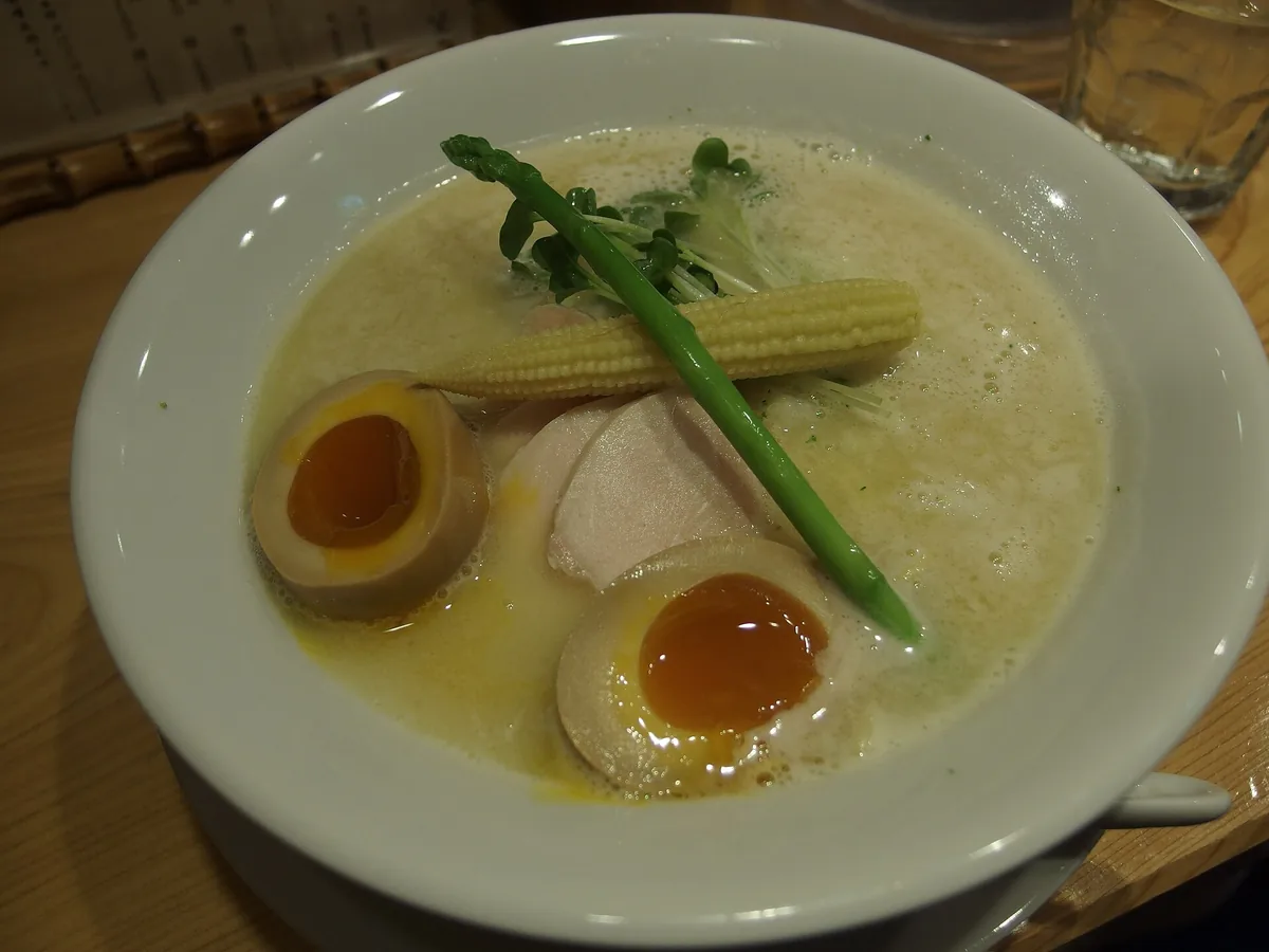 A ramen bowl in thick white broth with soft eggs, sliced chicken, asparagus and baby corn