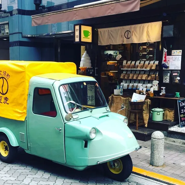 A pale green three wheeled truck parked in front of a shop with bag shelves and a cloth curtain