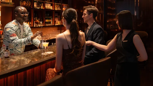 A bartender serving a cocktail to a couple seated at a marble counter, against shelves of bottles