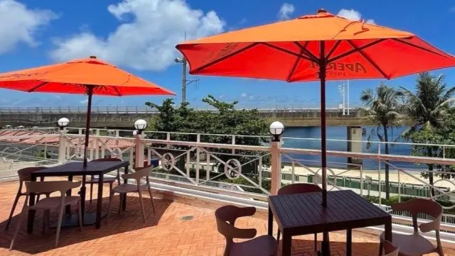 A terrace with orange parasols and dark tables, facing a bridge, sea and blue sky