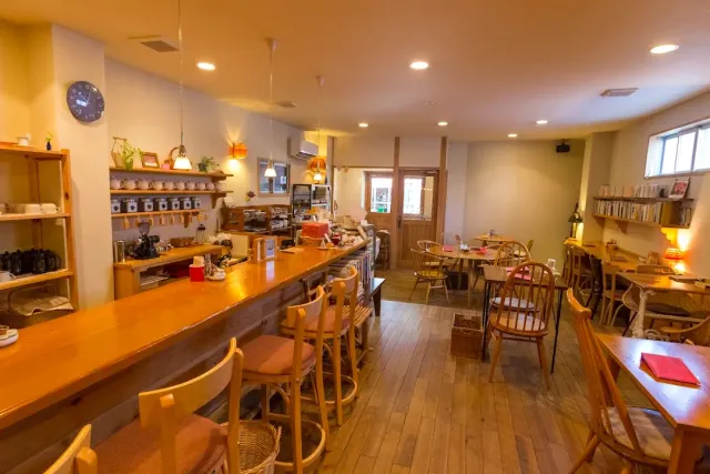 A long wooden counter with stools, shelves of cups, a blue clock and tables behind