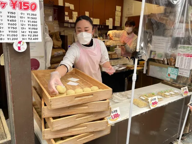A worker holding a wooden tray of greenish dough balls, in front of price signs in the shop