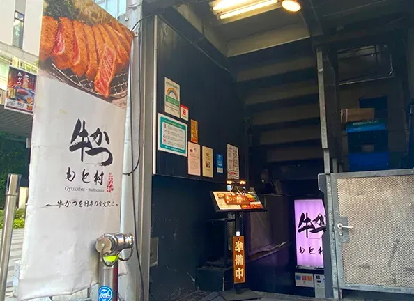 A narrow entrance with a banner showing slices of grilled meat, a lit purple sign and notices on the wall