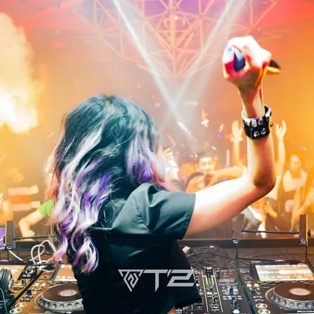 A female DJ raising her hand above the decks before a dancing crowd and stage lights