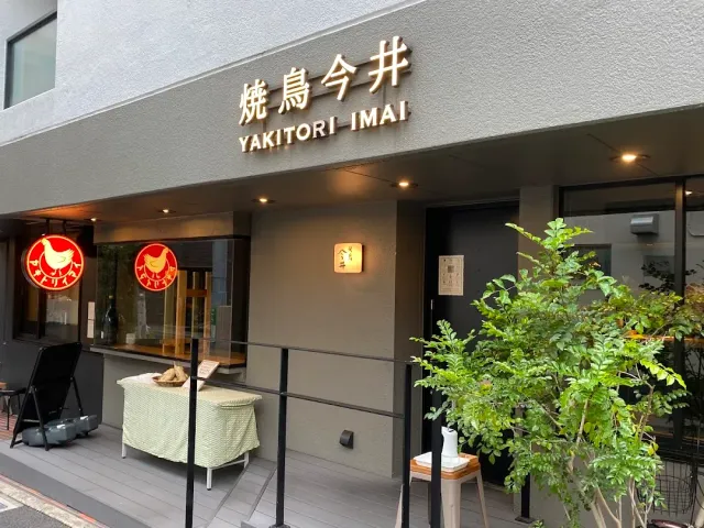 A pale frontage with raised lettering, two round red signs with a rooster and a low tree