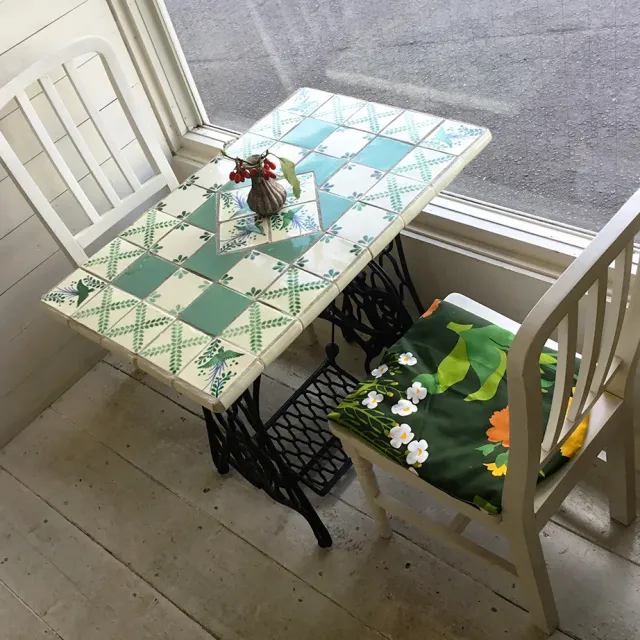 A table topped with green and white tiles between two white chairs, next to a window