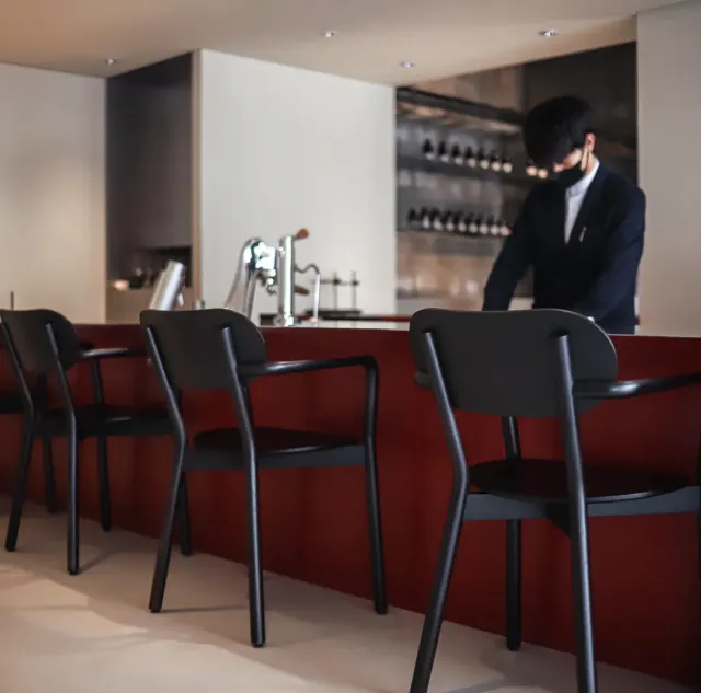 Black chairs facing a red counter, with a worker and a bottle shelf behind it