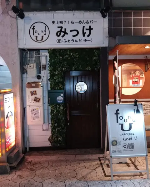 A narrow storefront in the evening with a wooden door, a plant wall and a standing sign on the pavement