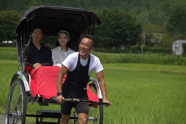 A smiling rickshaw puller draws a couple of passengers along green rice fields