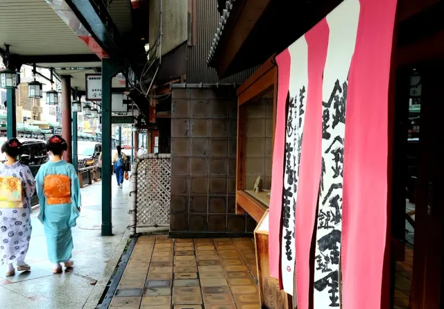 A covered street with pink and white cloth banners, and two women in kimono walking along it