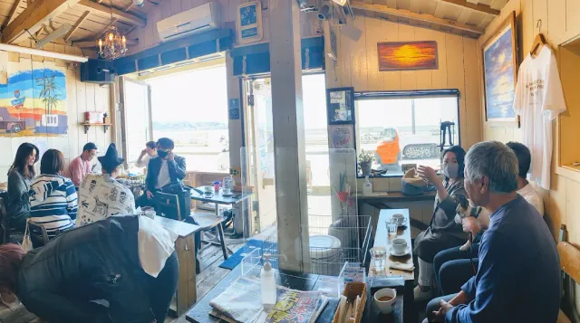 A small room full of guests with white wooden walls, pictures and the sea seen through the opening